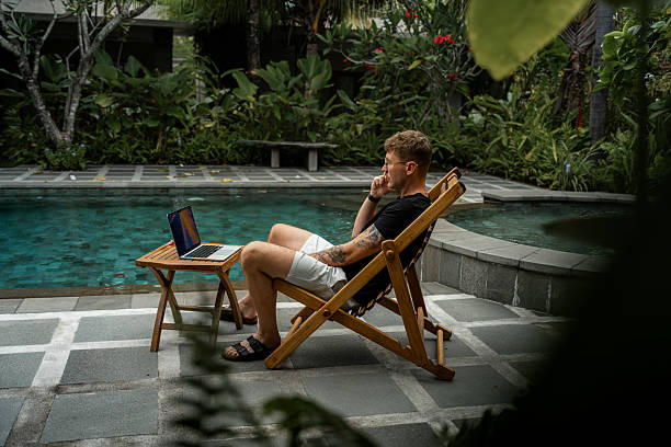Man speaks on phone while using laptop by swimming pool surrounded by a garden